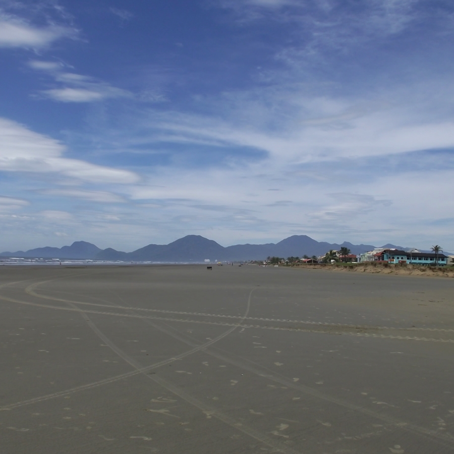 vista panorâmica de toda extensão da praia com morros e céu azul distantes ao fundo 